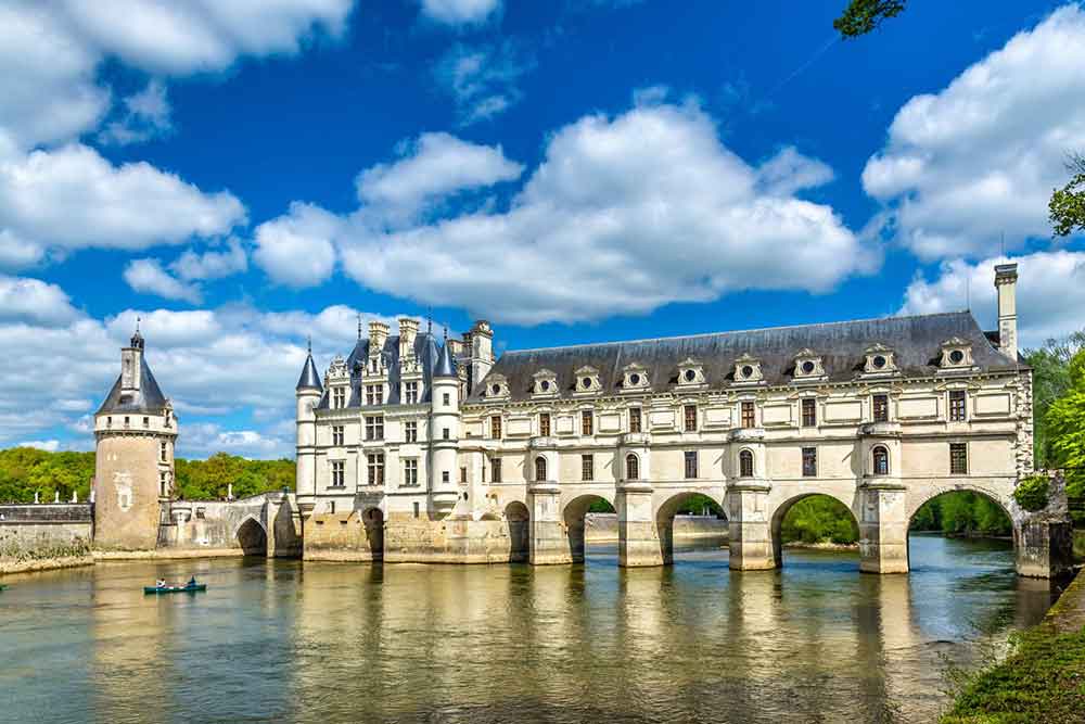 Château de Chenonceau, France, a fairytale castle, showcases elegant arches and a gray roof standing in the middle of the Cher River, under a clear, cloudy blue sky.