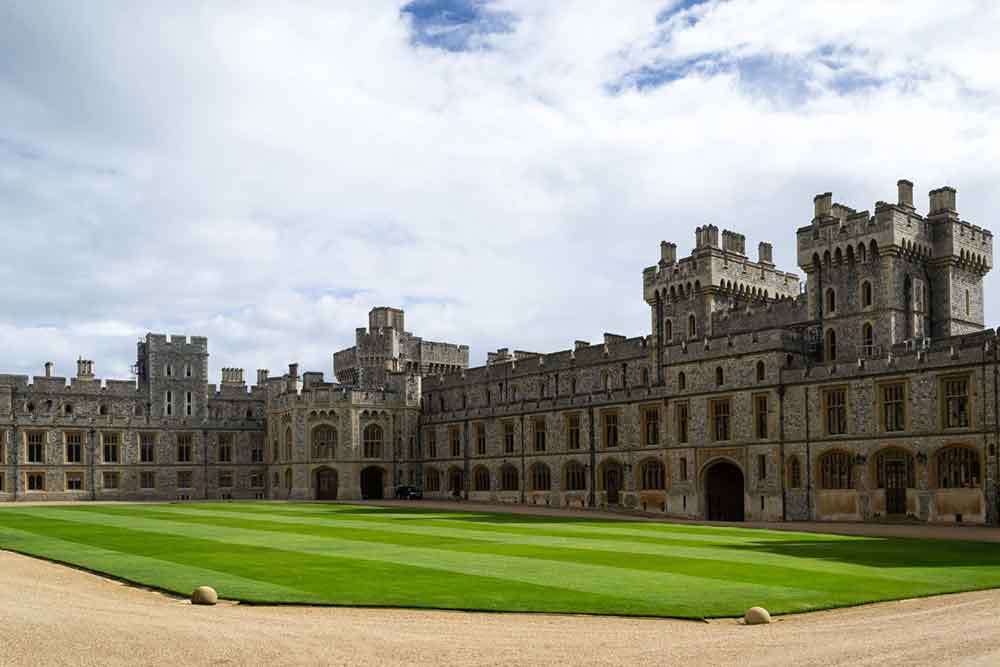 Windsor Castle, England features a massive stone walls, fortified towers and battlements with a green lawn in front of the castle