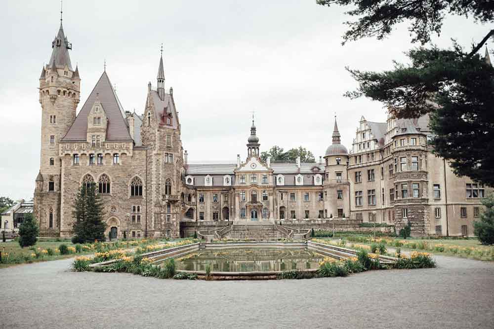 Moszna Castle in Poland, with its diverse architectural styles, multiple windows, and spires, accompanied by a pond in front of the castle and trees on the side