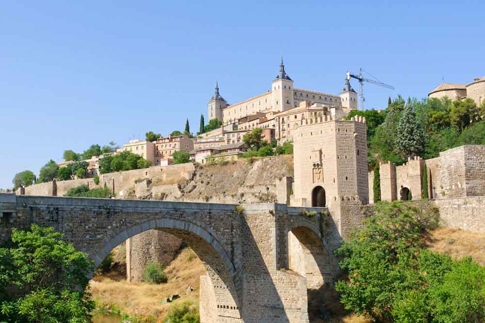The medieval Puente de Alcántara leading up to the historic skyline of Toledo, with its towers and stone walls rising above the gorge.