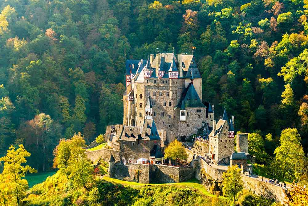 Eltz Castle in Germany features multiple towers, turrets, and gable roofs, with sandstone walls surrounded by a dense forest on a sunny day