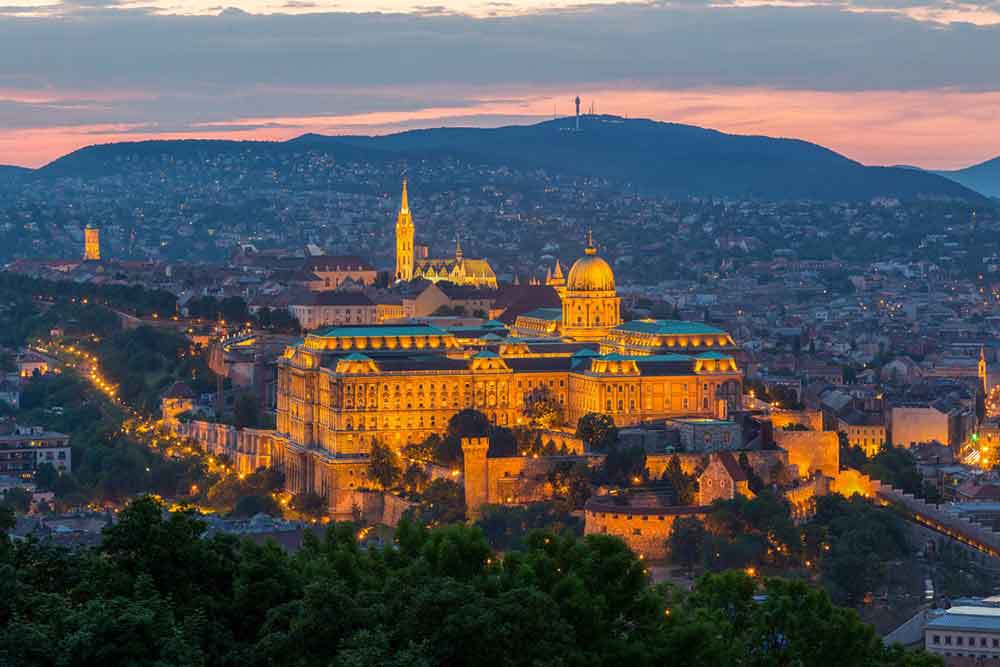 Buda Castle located in Hungary - the castle illuminated facade casts a warm glow light againts the night sky highlighting its dome, architectural designs and walls surrounded by cityscape and trees