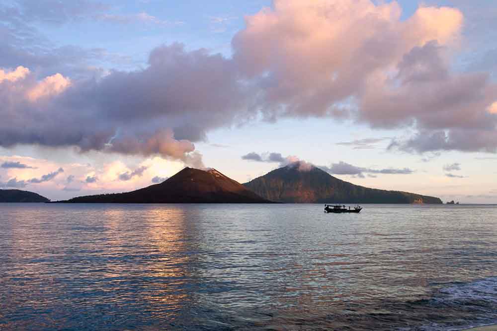 Mount Krakatoa, West Java featuring a volcanoes with white smoke rising from the sea ,set against the backdrop of a sunset and a boat nearby