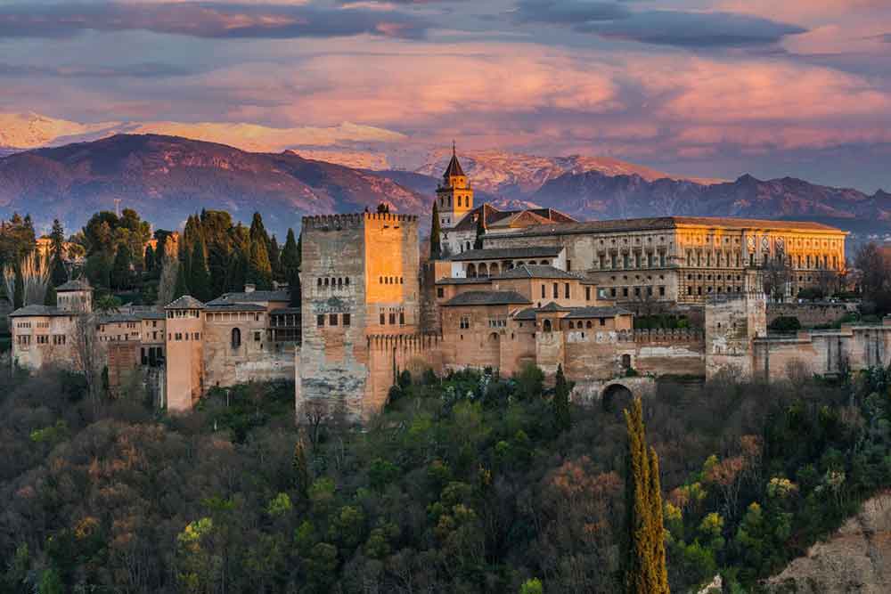 Alhambra in Spain features battlement high walls in a peach hue, surrounded by trees, and offers views of the mountains during sunset