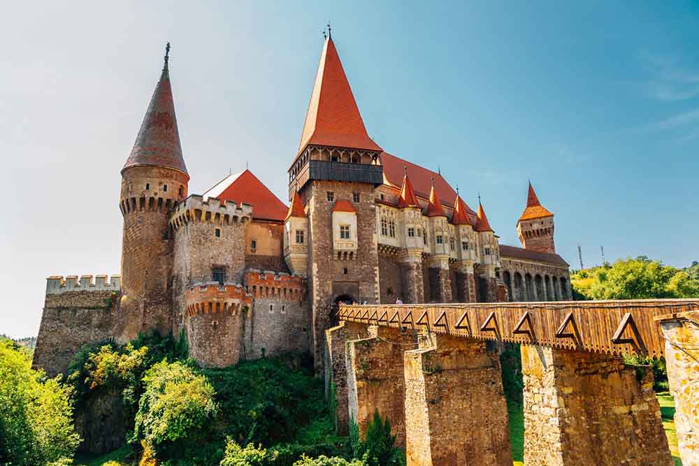 Corvin Castle, with its sand-brick walls and red turrets, greeted by a fortified entrance bridge under a clear blue sky