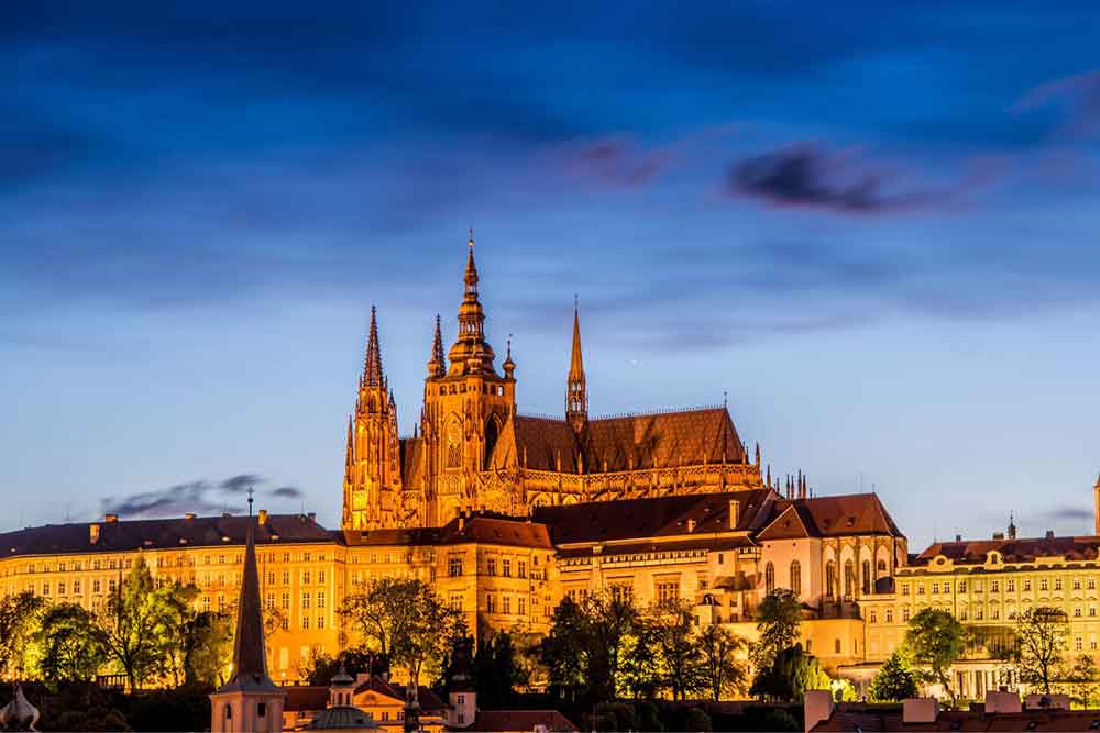 Prague Castle in the Czech Republic showcases spires, towers, and roofs that glow at night, surrounded by trees.