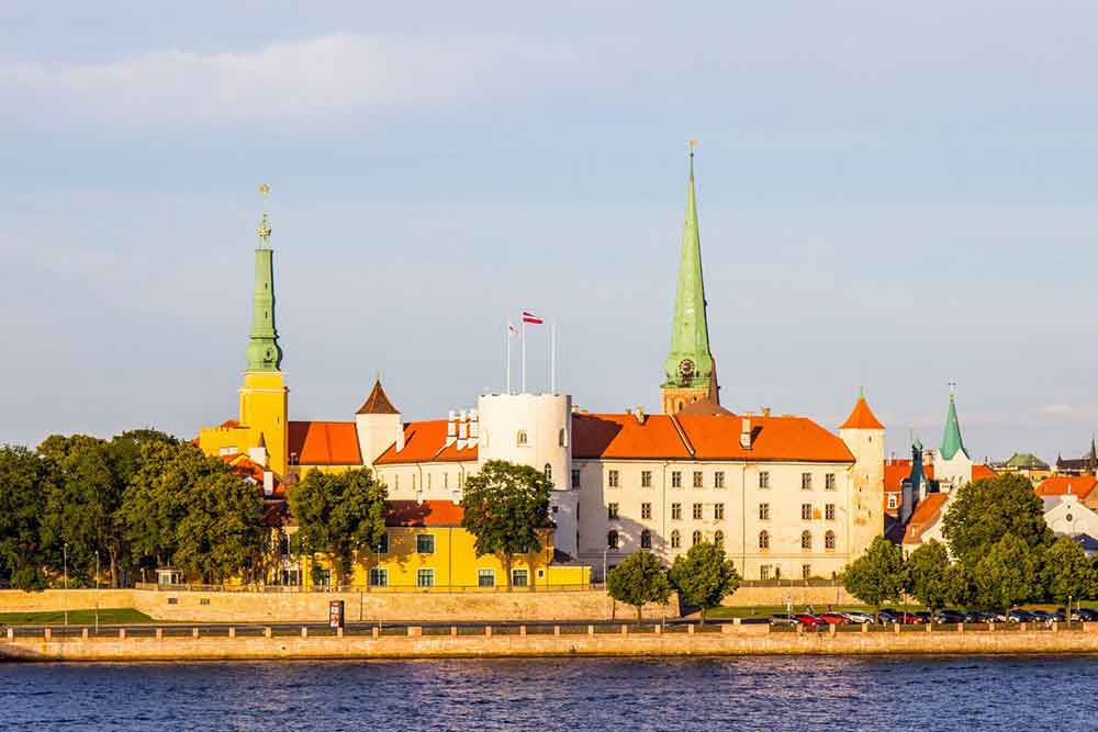 Riga Castle in Latvia features a red roof and white-yellow painted walls, along with two towers, surrounded by trees and water