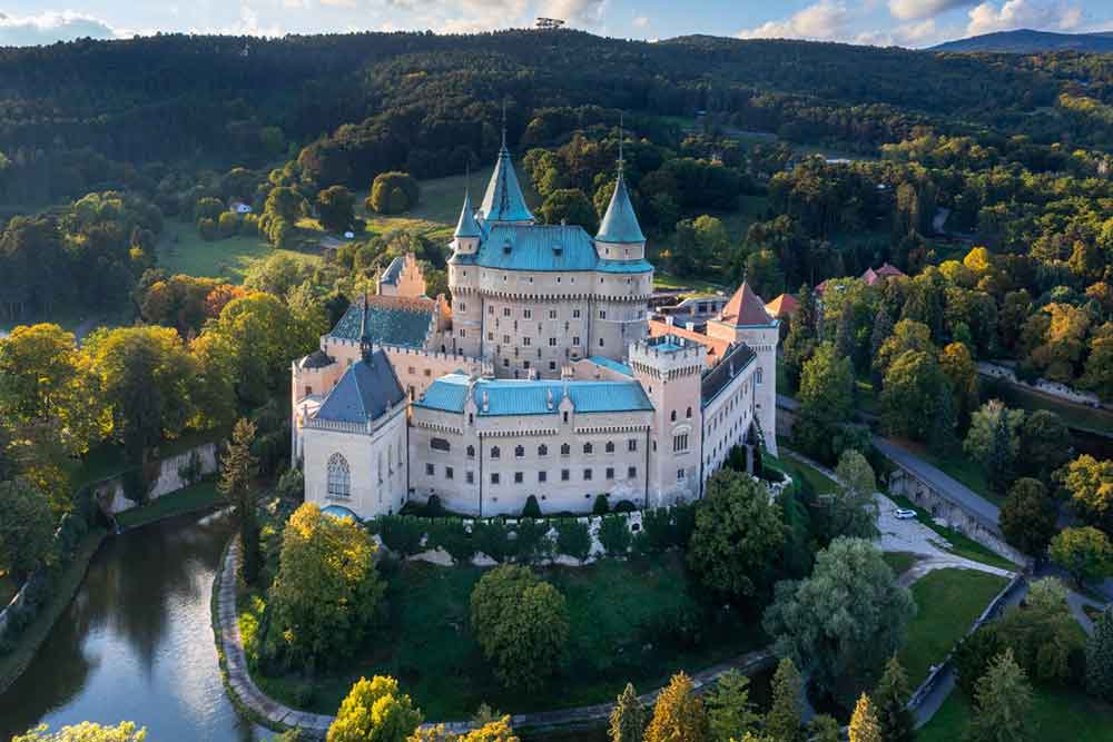 An aerial view of Bojnice Castle in Slovakia shows it perched on a hill, featuring a blue roof and high walls, all surrounded by lush trees