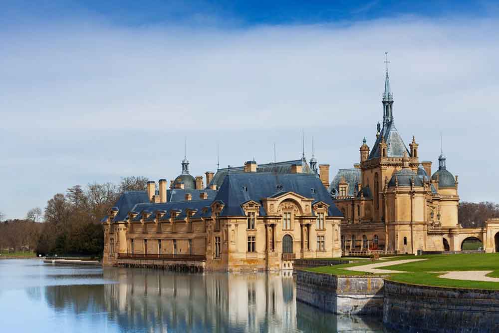 Domaine De Chantilly, France features a dark blue roof, spires and earthy walls surrounded by lake and manicured gardens