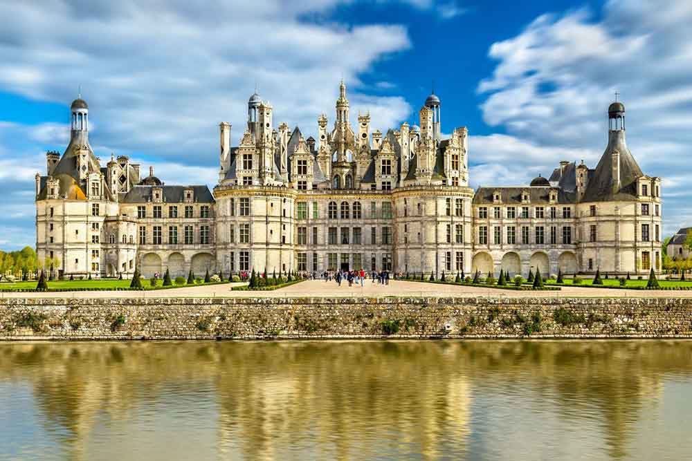 Château de Chambord in France, with its majestic structure featuring multiple windows, spires, and turrets, has a fairy-tale appearance, complemented by gardens and a river at the front