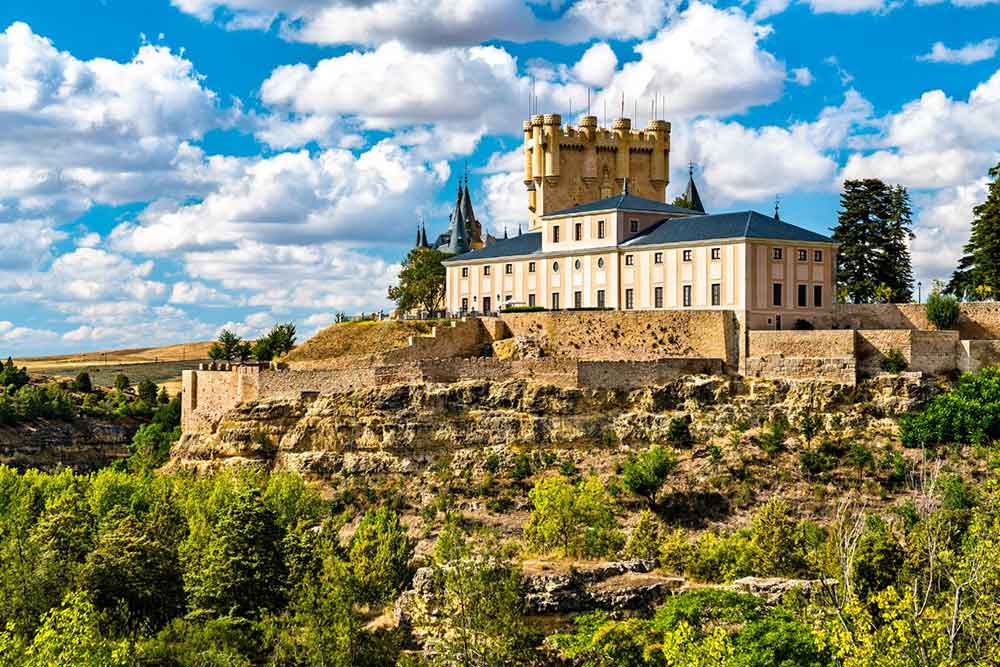 Alcazar of Segovia in Spain sits atop a rocky hill, featuring peach-white walls, a gray roof, and brown towers, all against a blue sky in the background