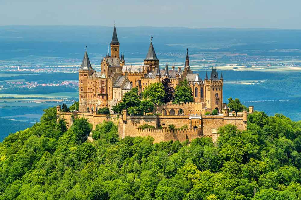 Hohenzollern Castle in Germany stands atop a hill, featuring a warm brown and golden hue - fairy-tale castle offers breathtaking views of the surrounding landscape and trees
