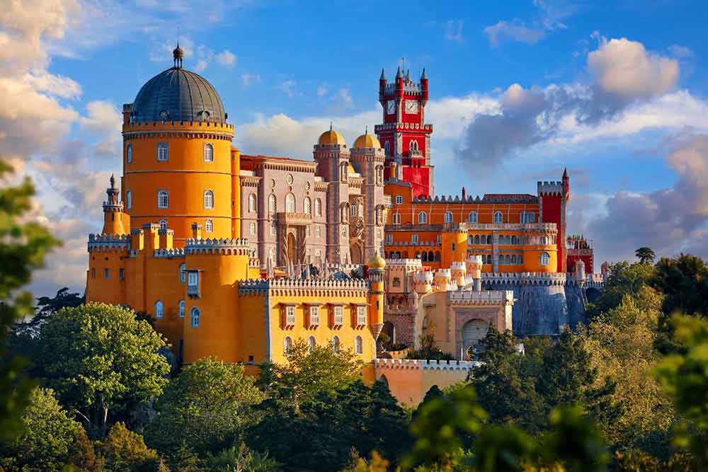 Pena Palace in Portugal with its yellow and red painted wall surrounded by trees in a clear blue sky