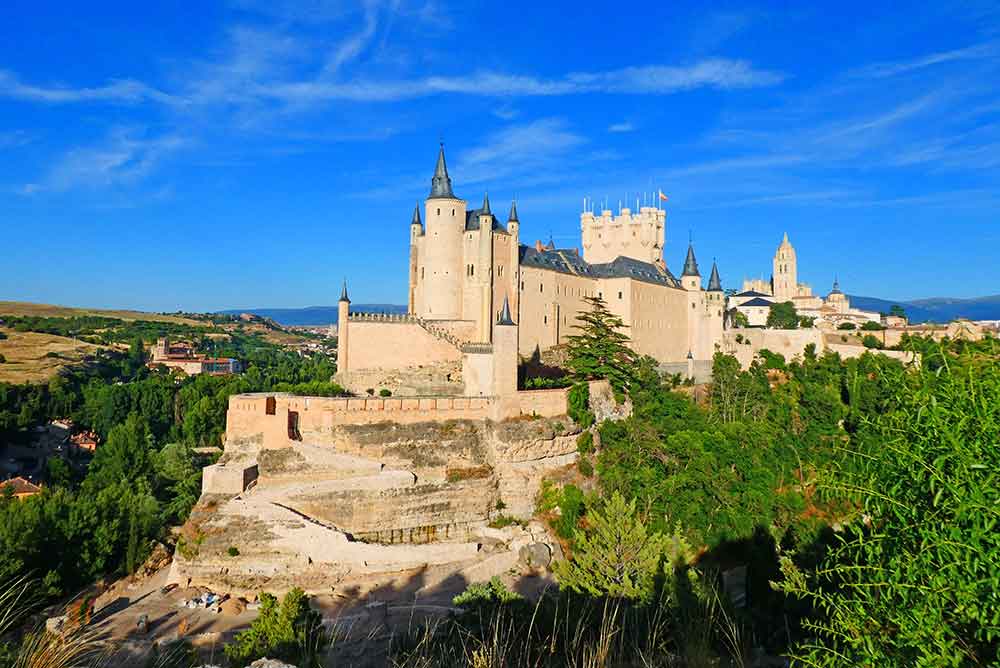 The Alcázar of Segovia perched on a rocky hilltop, its towers rising above the surrounding greenery and panoramic landscape.