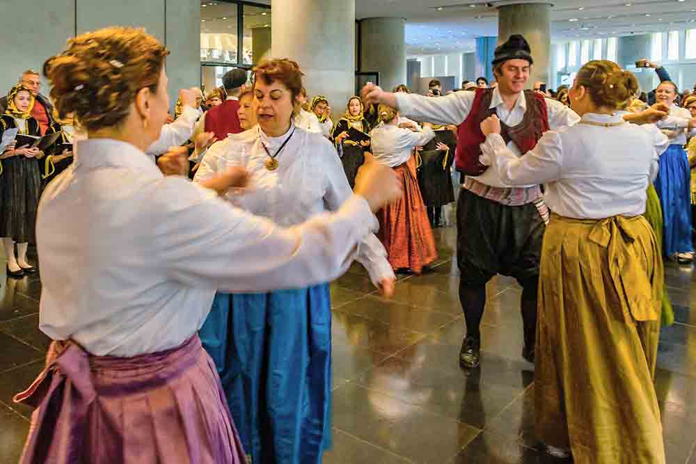 Greek dancers wearing traditional outfits and dancing with people in the background singing