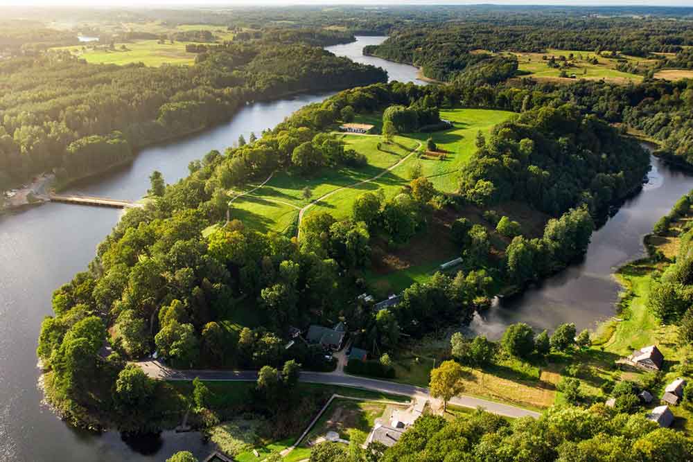 An aerial view of Dubingiai Castle remains surrounded by a lake, forested trees, and green lawns.