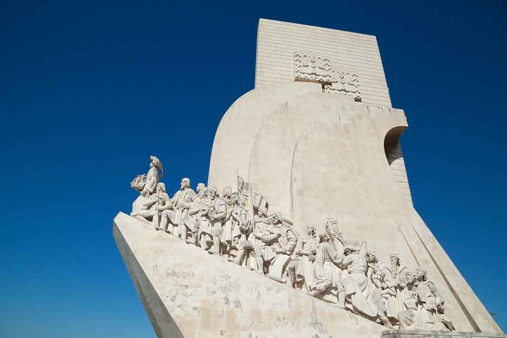 The Monument of Discoveries in Lisbon sits on an enormous marble mosaic and features carved figures against a clear blue sky.