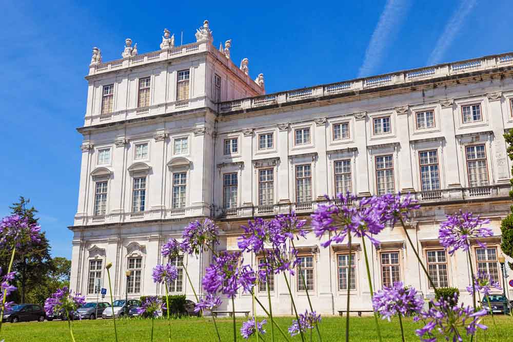 Ajuda Palace in Lisbon features a long building with multiple windows and carved statues at the top, all set against a backdrop of violet flowers in the garden.