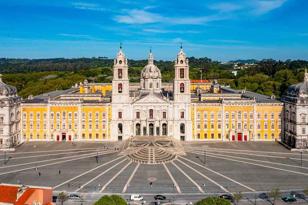 Palácio Nacional de Mafra in Lisbon is a large palace painted in yellow and cream, with three towers at the center and spacious grounds.