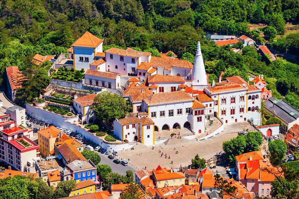 An aerial view of the Palácio Nacional in Sintra features multiple buildings with red-orange roofs, surrounded by trees and houses.
