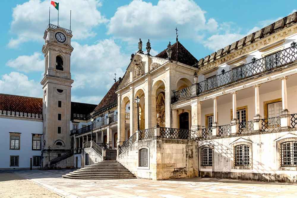 Coimbra University, one of the oldest universities in the world, features a staircase at the entrance with multiple columns, cream-white walls, and a clock tower that stands tall and proud.