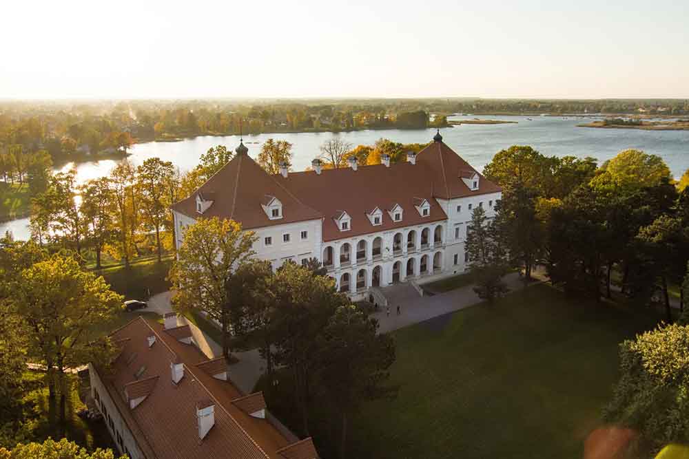 An aerial view of Birzai Castle in Lithuania features red roofs and gray-white stone walls, surrounded by a landscape of trees and a lake.