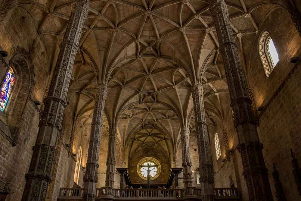 Interior view of Jeronimos Monastery showcases its spacious and high ceiling with intricate stone carvings on the columns, arches, and walls.