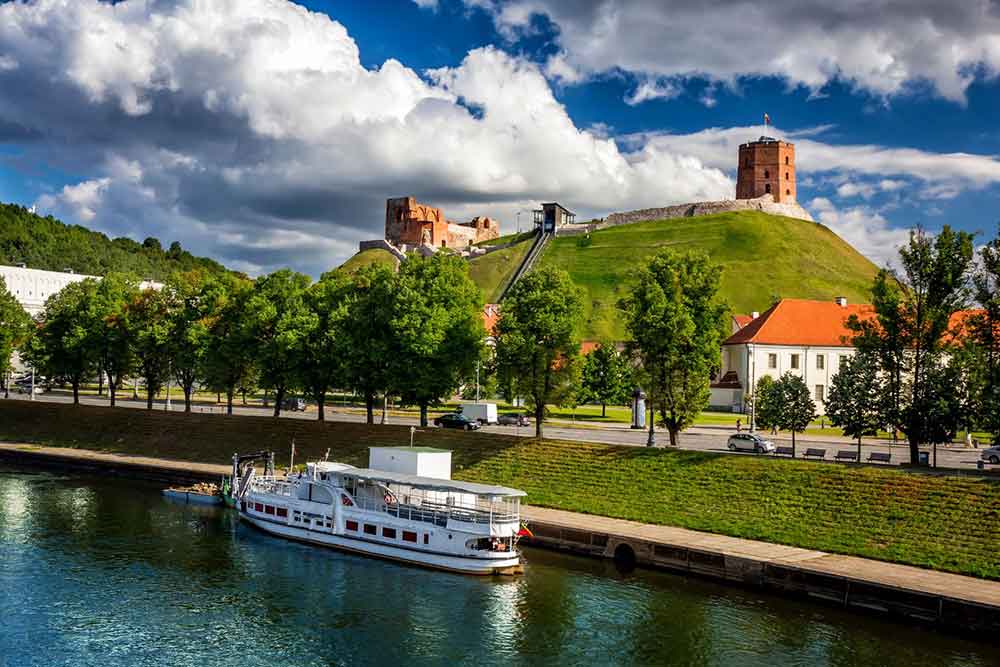 Vilnius Castle in Lithuania sits atop a hill, featuring a red-brick tower that overlooks the city, trees, and a lake.