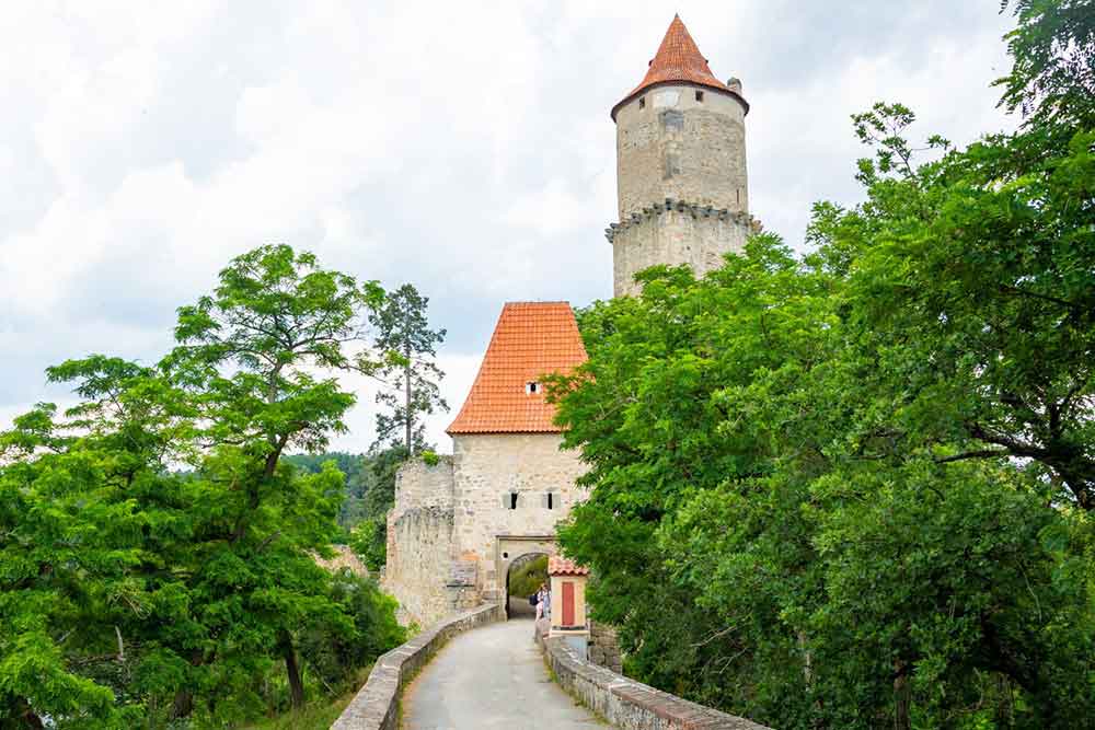 The entrance to Castle Zvíkov with a bridge going though a gate and a tower in the background surrounded by trees 