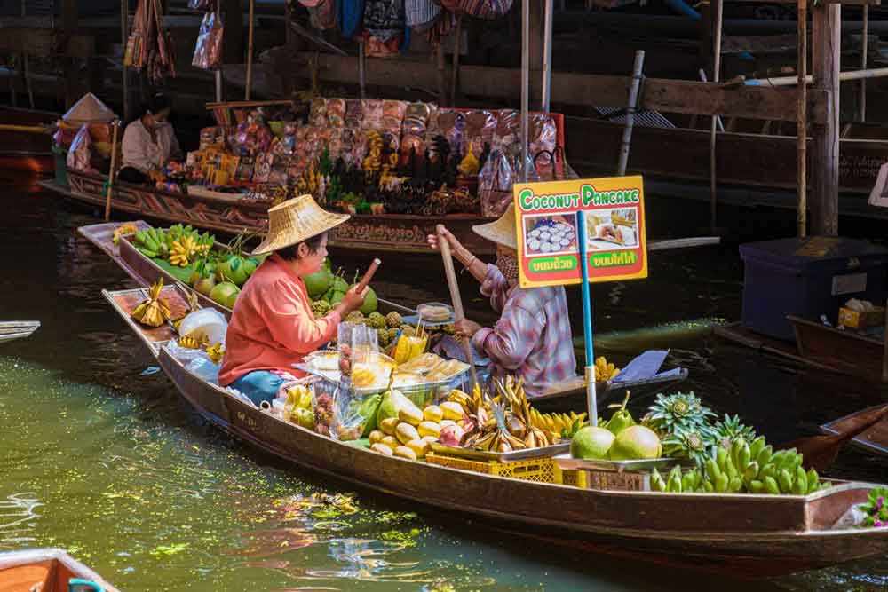 Two vendors selling fruits and vegetables from their boats at the Damnoen Saduak Floating Market in Thailand.