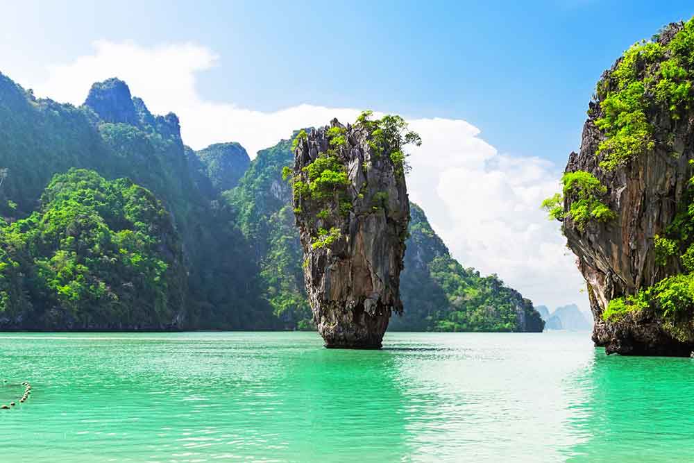 James Bond Island in Phang Nga Bay features a vertical rock formation standing at the middle of the sea water, with large rock formations in the background.