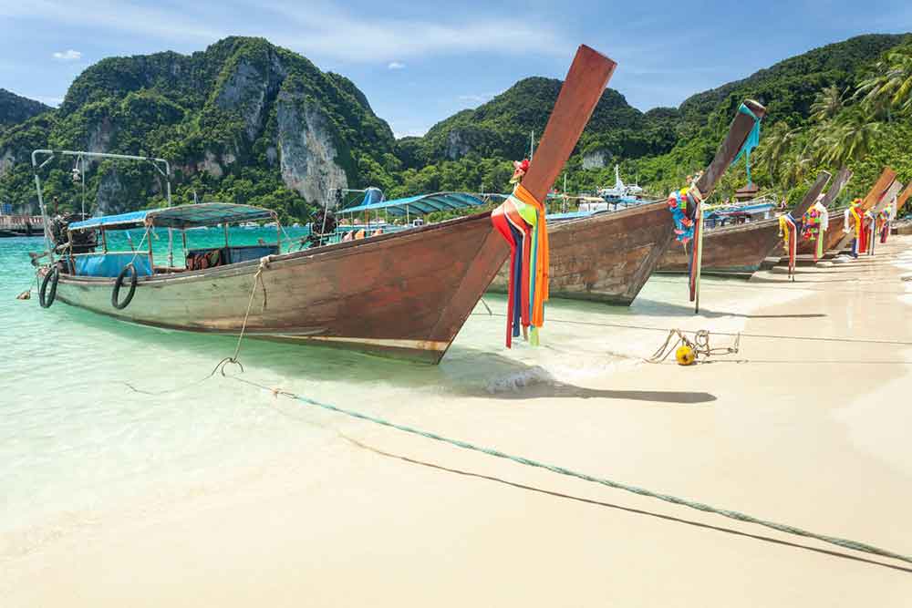 Longtail boats parked at the shoreline on Koh Phi Phi Island with mountains in the background, all under a clear blue sky.