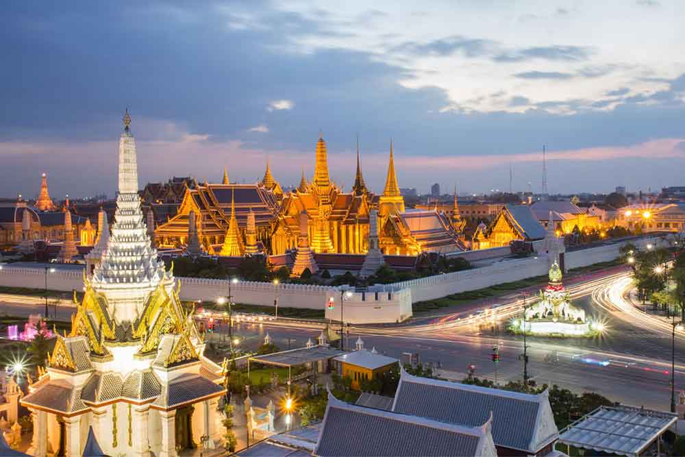 The Grand Palace and Wat Phra Kaew illuminated by lights at night, with bustling streets and a highway in Thailand.