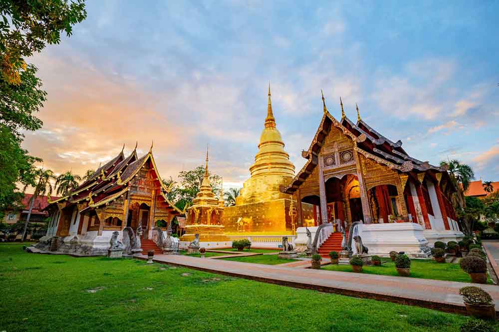 Wat Phra Sing Temple in Thailand features a large golden prang with spires at the center, flanked by two temples on the sides, all with multi-tiered red roofs, under a clear blue sky.