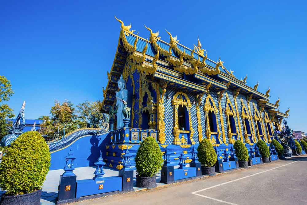 Wat Rong Sua Ten, also known as The Blue Temple in Thailand, features an all-blue and gold structure with a statue standing at the entrance, set against a clear blue sky.