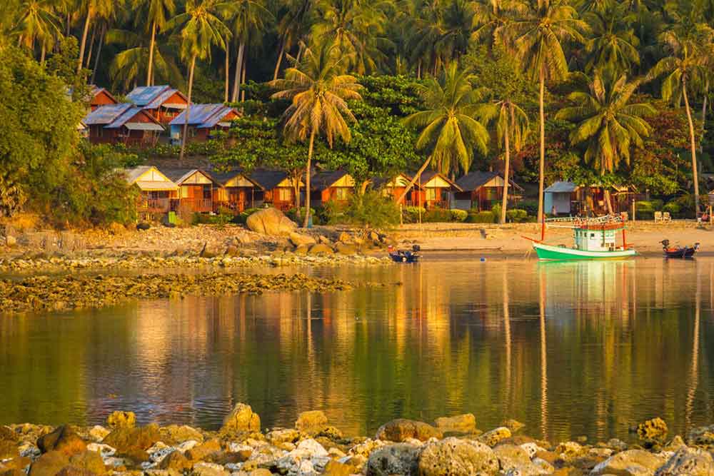 An amazing sunset in Ko Pha Ngan, Thailand unfolds with houses, coconut trees, and the calm water with small boat