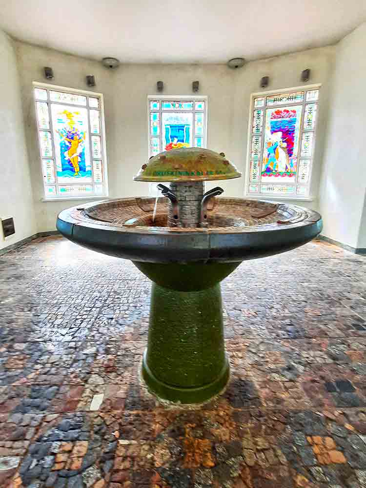 Druskininkai public water fountain an ornate fountain with fout taps spouting mineral water inside a room with stained glass windows in the background. 
