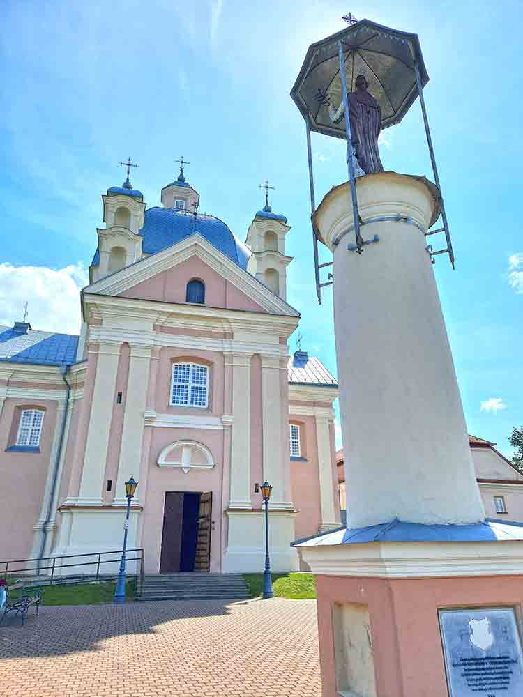 Pink and light grey Liškiava Monastery facade with statue int he foreground of a man under an umbrella with blue skies and a few clouds in the background.