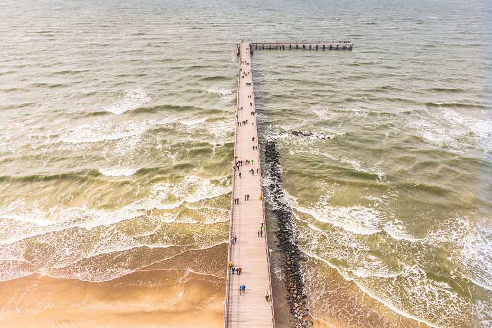 An inverted 'L' shaped pier justting out to sea. There are a few white waves and the water is greenish with some sand at the forground of the picture.
