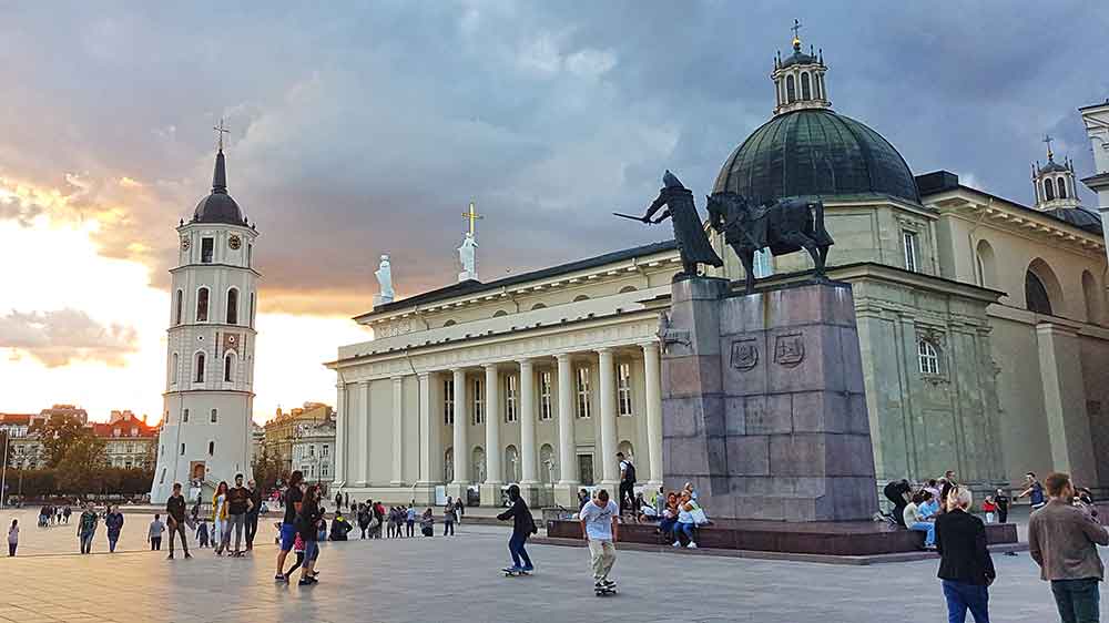 The old town of Vilnius with a tower, statue of a horse on a plinth and ornate building with an open plaza and lots of people. The sky has one giant dark grey cloud but the sun i the background.