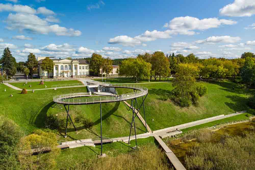 A spiral shaped bridge set in the bank of the lake with an ornate buidling behind it. There is green grass and trees with a blue sky.