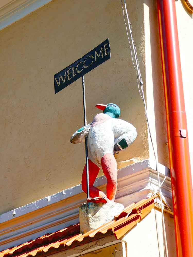 Užupis artwork featuring a duck stood upright with one hand onhis hip and the otherholding a sign that says 'welcome'. He is stood on a roof of a building. 
