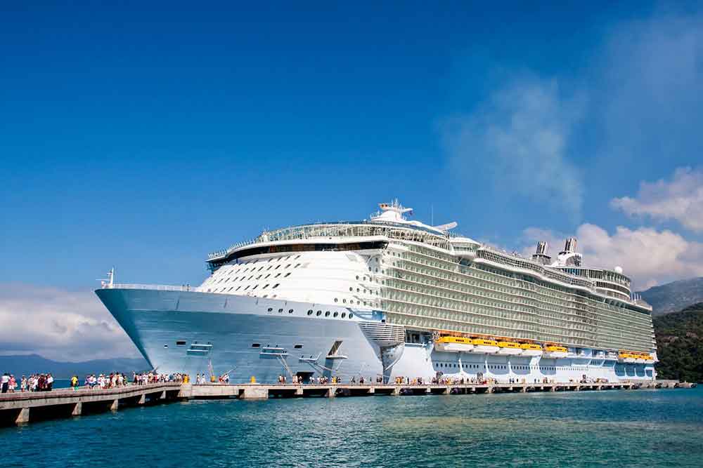 a large white cruise ship sat in dock against a pier with people on the pier. There is blue sky and blue water.