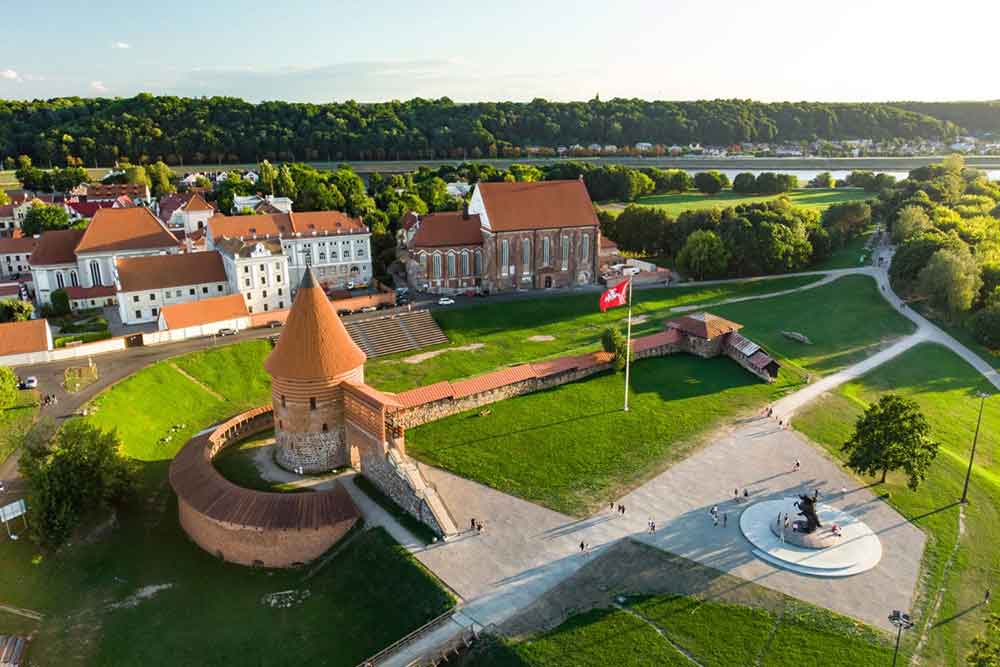 Kaunus castle with red bricks and red roof tiles. There are only a few walls remaining but also a rotund tower with a conical roof with a lower circular bulding. 