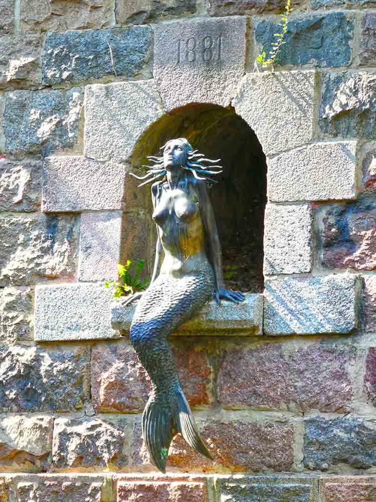 The bronze statue called mermaid of Užupis sitting inside a little alcove in a wall. She is looking up. The wall says 1881 above where she is sitting.