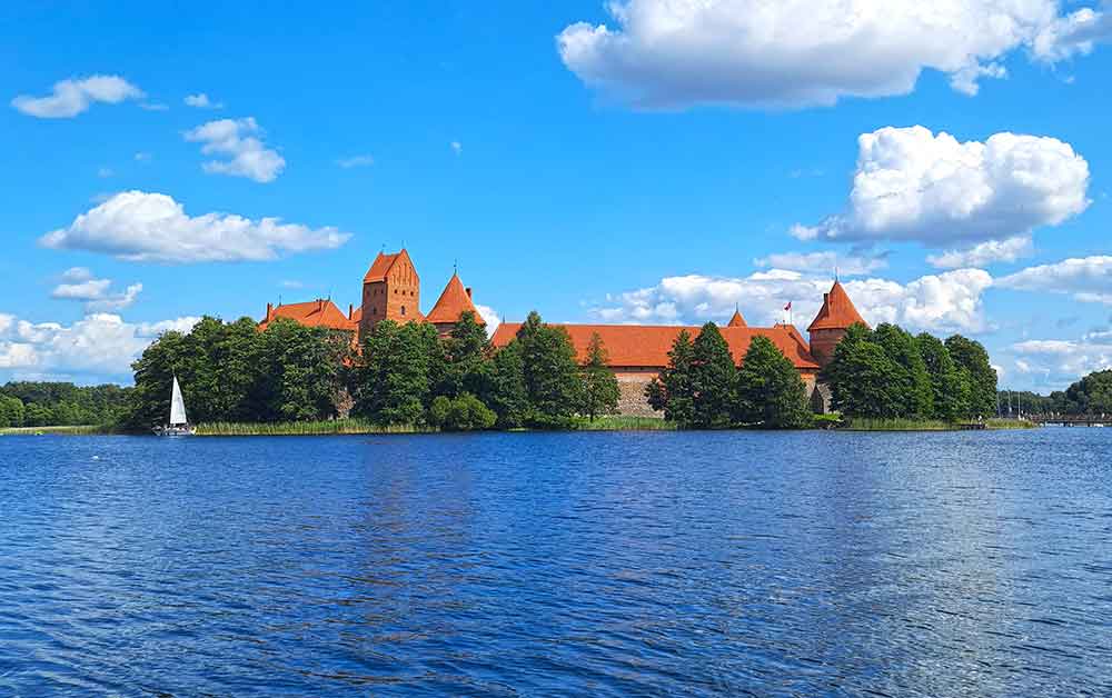Trakai castle - with red bricks and red roof situated on an island with lots of green trees. The water and sky is blue. There are some fluffy white clouds in the sky. 