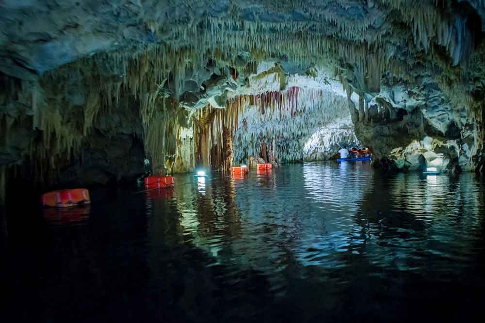 Is Peloponnese Worth Visiting? 15 Reasons to Visit the Peloponnese Region, Greece 6 Diros Cave showing stalacties hanging from the ceiling and a river flowing through it. There is a small boat carrying people in the background.