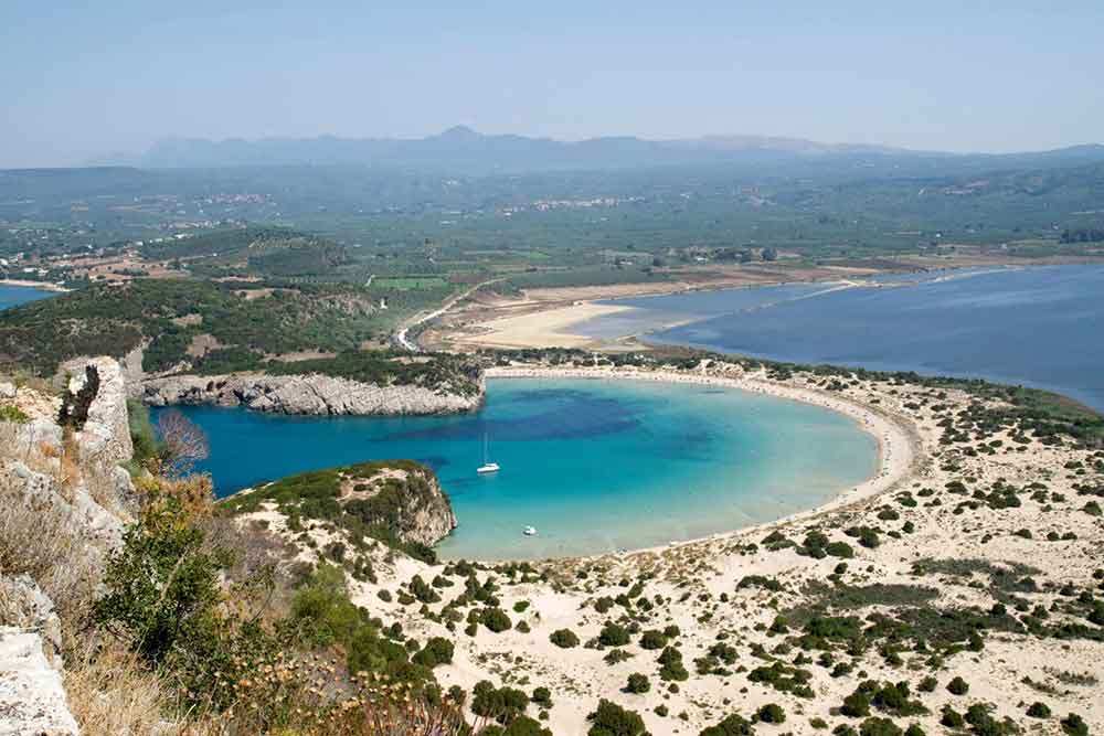 Is Peloponnese Worth Visiting? 15 Reasons to Visit the Peloponnese Region, Greece 8 The curved bay of Voidokoilia beach with an arced shaped blue sea and white sand with small green shrubs.
