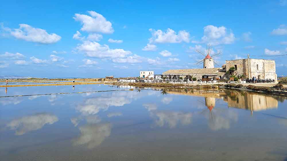Trapani and Paceo Salt Pans with a windmill and light brick buildings. These are reflected in the water.