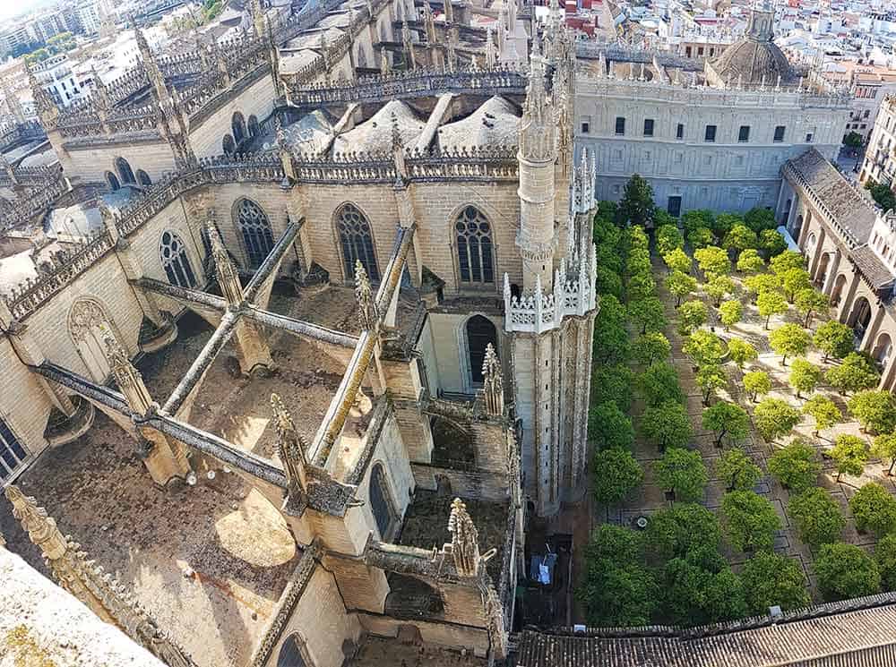 View looking down from the Giralda Tower in Seville. There is an orangery with rows of orange trees laid out in a plaza, and the Gothic roof of the cathedral.
